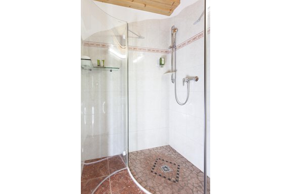 Eine moderne Dusche mit Glaswänden und einem Bodeneinlauf. Die Wände sind hell gefliest und es gibt einen Duschkopf sowie Ablagen für Pflegeprodukte. - Hotel Stern - Längenfeld