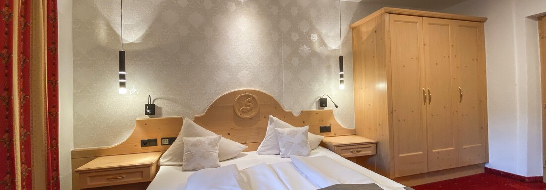 Ein gemütliches Hotelzimmer mit einem Doppelbett, zwei Kopfkissen und Holzdetails. Die Wände sind in sanften Farben gehalten, und es gibt einen großen Kleiderschrank. - Hotel Stern - Längenfeld