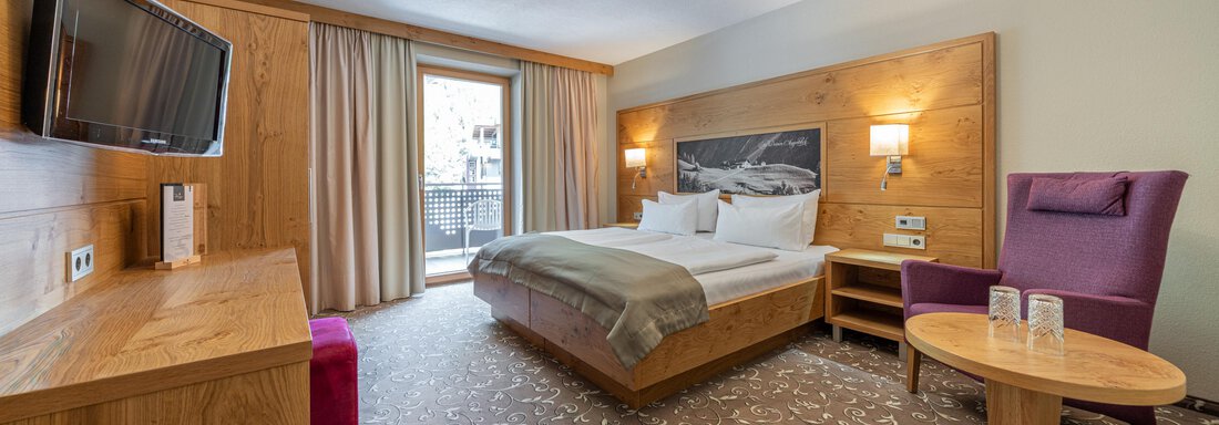 web-stern-zimmer-326-1016 Ein modernes Hotelzimmer mit einem Doppelbett und einem großen Fenster. Es gibt Holzdetails, einen Fernseher und eine gemütliche Sitzecke. - Hotel Stern - Längenfeld