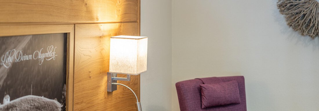 Ein modernes Zimmer mit einem bequemen lila Sessel und warmem Holzinterieur. An der Wand hängt ein kunstvolles Licht und ein Bild einer Landschaft. - Hotel Stern - Längenfeld