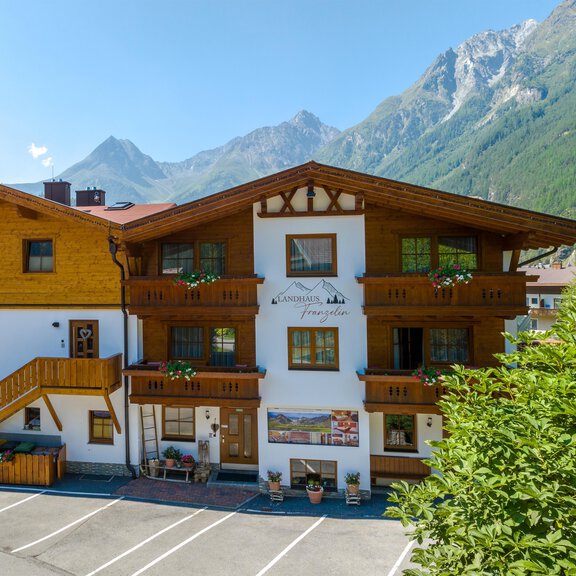 Ein charmantes Fachwerkhaus mit Holzverkleidung in den Alpen. Im Hintergrund sind beeindruckende Berggipfel und ein blauer Himmel zu sehen. - Landhaus Franzelin - Längenfeld