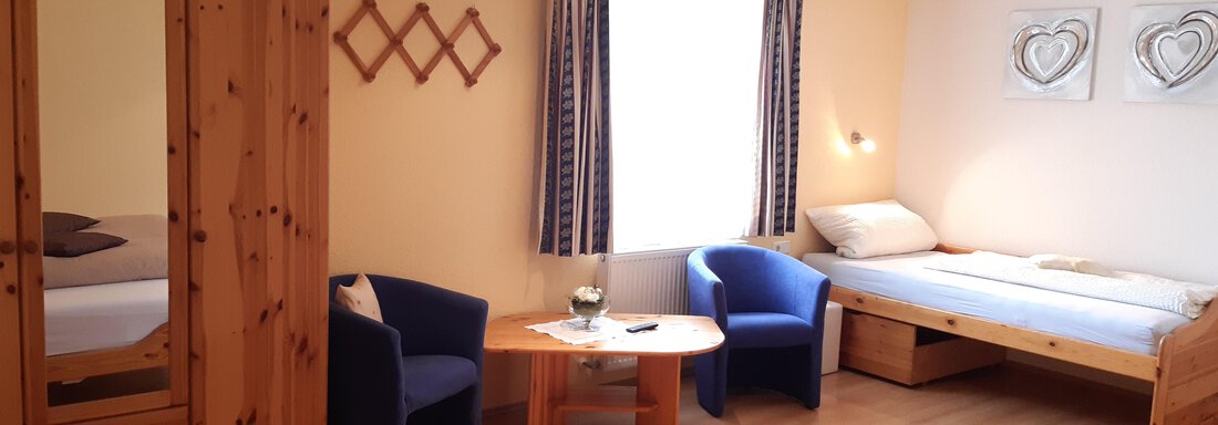 A cozy room with two single beds and a table. The decor is bright and friendly, with blue chairs and wooden shelves. - Landhaus Ötztalblick - Längenfeld