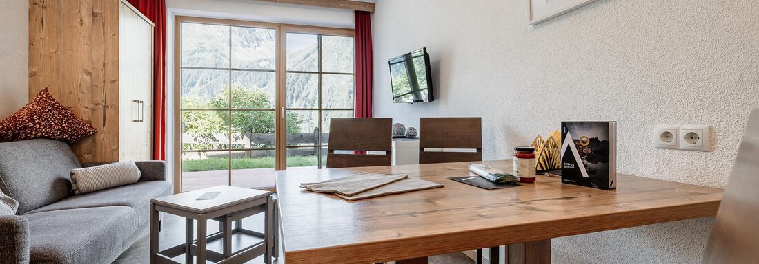 A modern living and dining room with a cozy sofa and a wooden table. Large windows offer a view of the garden and let in plenty of light. - MYALPS Himmelreich - Längenfeld