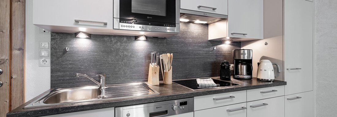 A modern kitchen with white cabinets and a dark gray countertop. It features a stove, a microwave, and appliances for coffee preparation. - MYALPS Himmelreich - Längenfeld