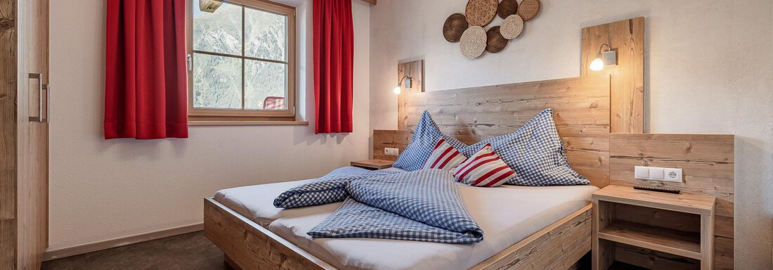 A cozy bedroom with a wooden bed and colorful cushions. Red curtains and a window area create an inviting atmosphere. - MYALPS Himmelreich - Längenfeld