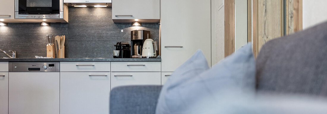 A modern kitchen with bright cabinets and appliances that is well-equipped. In the foreground, a gray sofa with cushions is visible. - MYALPS Himmelreich - Längenfeld