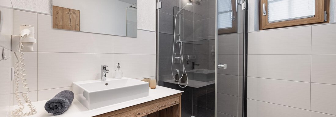 A modern bathroom with a glass shower and a sink on a wooden dresser. Bright tiles and windows create a friendly atmosphere. - MYALPS Himmelreich - Längenfeld