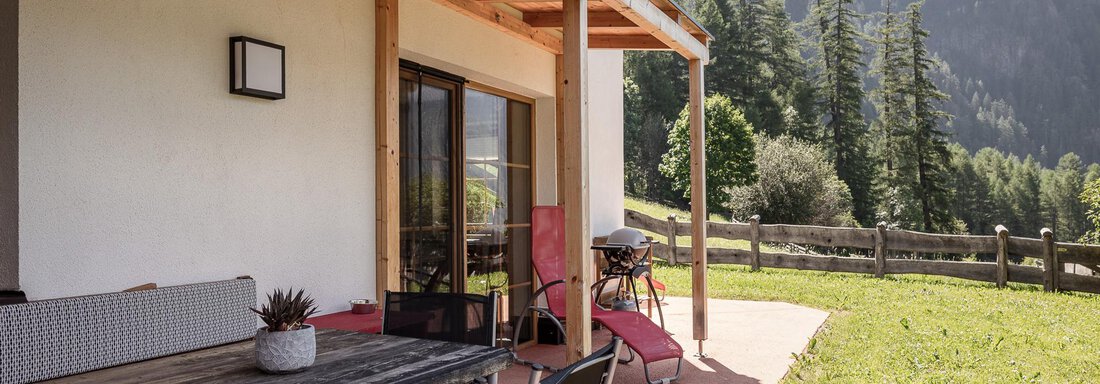 A cozy terrace with a wooden table and chairs, surrounded by green grass and mountains in the background. The sun is shining on the relaxing surroundings. - MYALPS Himmelreich - Längenfeld