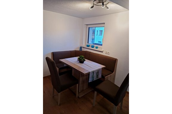 Ein gemütlicher Essbereich mit einer L-förmigen Bank und einem Tisch in Holzoptik. Im Hintergrund befindet sich ein Fenster mit blauen Dekorationen. - Apart Stefan - Niederthai