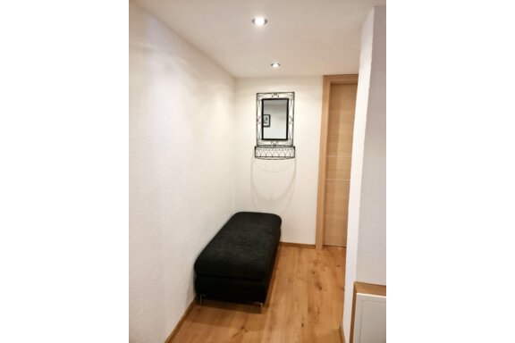Ein schmaler Flur mit einem schwarzen Sitzplatz und einem dekorativen Spiegel an der Wand. Der Boden ist aus Holz und die Wände sind hell gestrichen. - Apart Stefan - Niederthai