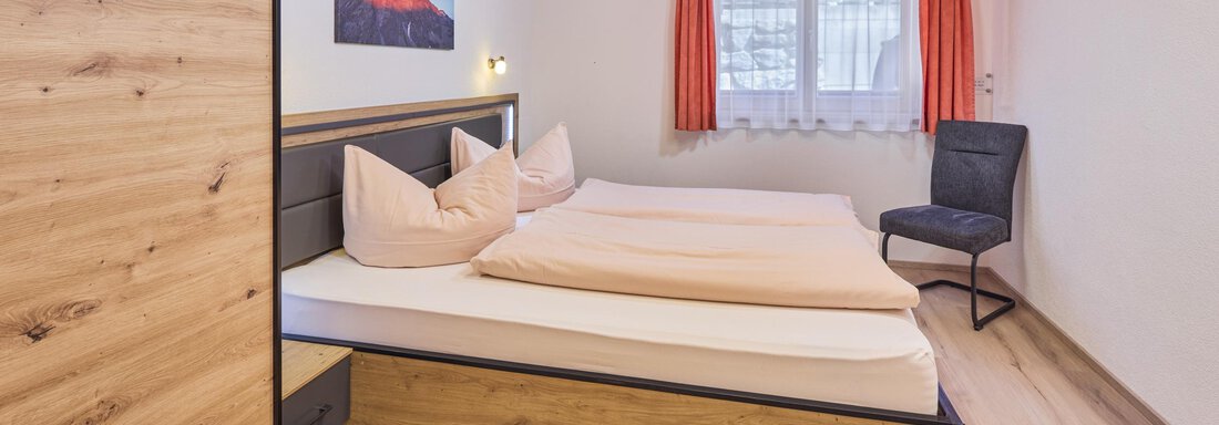 A cozy bedroom with a double bed and a chair. The walls are light and there is a window with curtains. - Appartement Lea - Niederthai