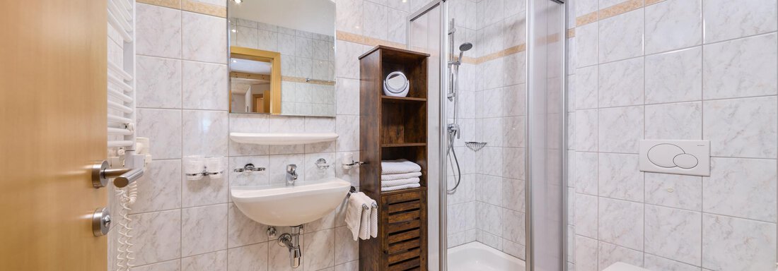 A modern bathroom with a shower, a sink, and a toilet. The walls are tiled and there are wooden shelves for towels. - Appartement Lea - Niederthai