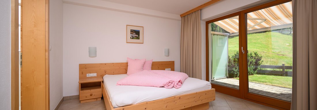 Photo 1 of the two bedrooms with a wooden bed and wardrobe. Large windows offer a view of the outdoor area and provide plenty of daylight. - Delano Appartements - Niederthai