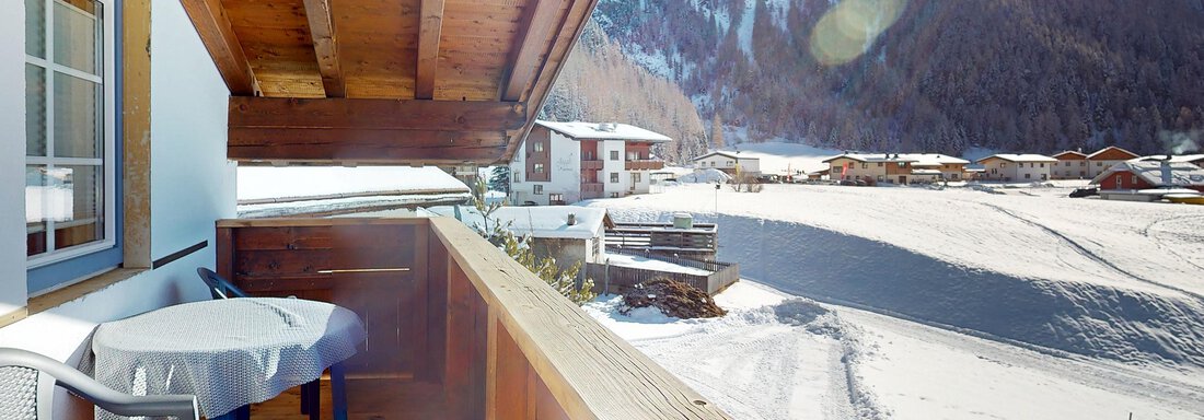 Ein Balkon mit einem Tisch und Stühlen, von dem aus man die verschneite Landschaft sehen kann. Im Hintergrund sind Berge und Häuser zu erkennen. - Gipfelblick Appartements - Niederthai
