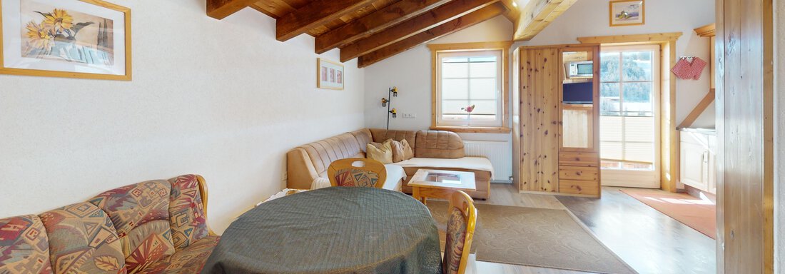 Ein gemütlicher Wohnraum mit Holzdecke, eingerichtet mit einem Tisch und einer Couch. Durch das Fenster fällt helles Licht auf den Raum. - Gipfelblick Appartements - Niederthai