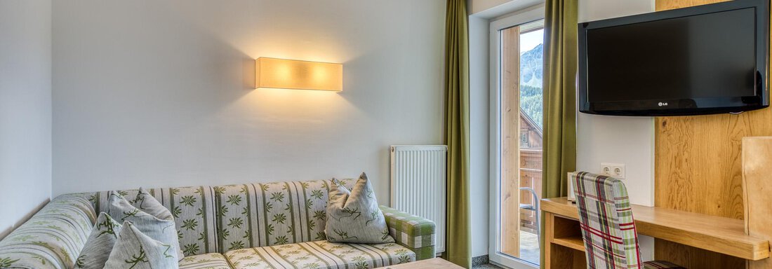 Ein gemütliches Wohnzimmer mit einem Sofa, einem Holzschreibtisch und einem Fernseher. Die hellen Vorhänge und das Fenster sorgen für eine freundliche Atmosphäre. - Hotel Falknerhof, am Ursprung - Niederthai