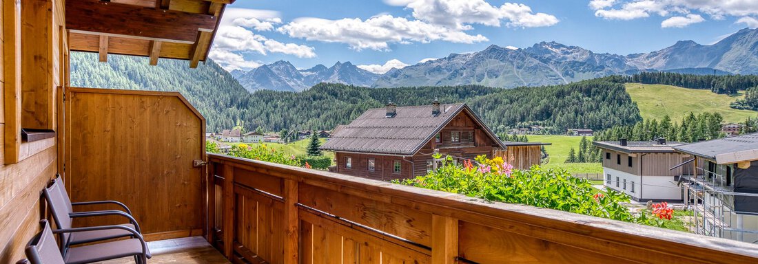 Ein Balkon mit Holzgeländer, von dem aus man auf die Berge und saftig grüne Wiesen blicken kann. Die Aussicht zeigt eine ruhige Landschaft unter blauen Himmel und weißen Wolken. - Hotel Falknerhof, am Ursprung - Niederthai