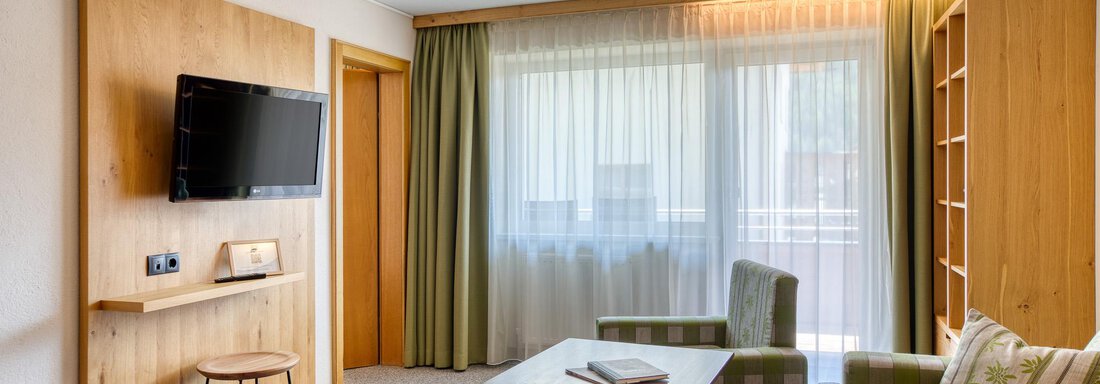 Ein gemütliches Wohnzimmer mit einem Fernseher, einem Tisch und bequemen Sitzmöbeln. Große Fenster lassen viel Licht herein und bieten einen Blick nach draußen. - Hotel Falknerhof, am Ursprung - Niederthai