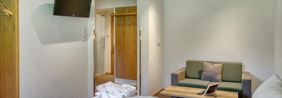 Ein modernes Hotelzimmer mit einem großen Bett, einem Sofa und einem Fernseher. Die Wände sind hell, und es gibt Holzdetails im Raum. - Hotel Falknerhof, am Ursprung - Niederthai
