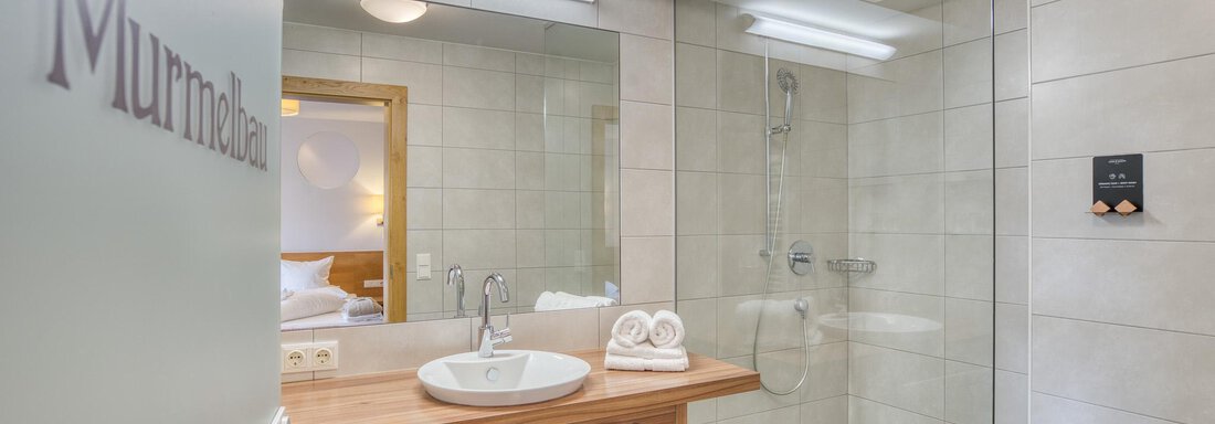 Ein modernes Badezimmer mit einem großen Spiegel und einer Glasdusche. Der Waschtisch aus Holz hat Handtücher und einen Korb mit weiteren Utensilien. - Hotel Falknerhof, am Ursprung - Niederthai