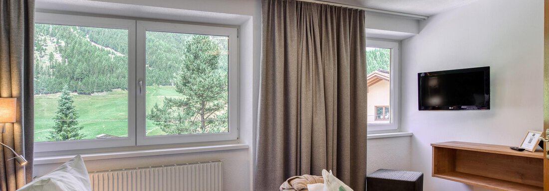 Ein helles Schlafzimmer mit Fenstern, die einen Blick auf eine grüne Landschaft bieten. Im Raum befinden sich ein Bett und ein Fernseher an der Wand. - Hotel Falknerhof, am Ursprung - Niederthai