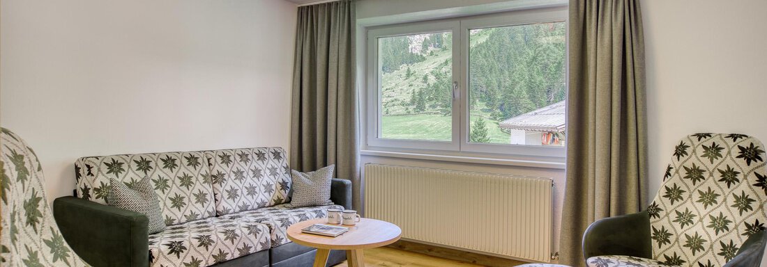Ein modernes Wohnzimmer mit einem Sofa und zwei Sessel, die ein florales Muster haben. Große Fenster bieten einen Blick auf die grüne Landschaft draußen. - Hotel Falknerhof, am Ursprung - Niederthai