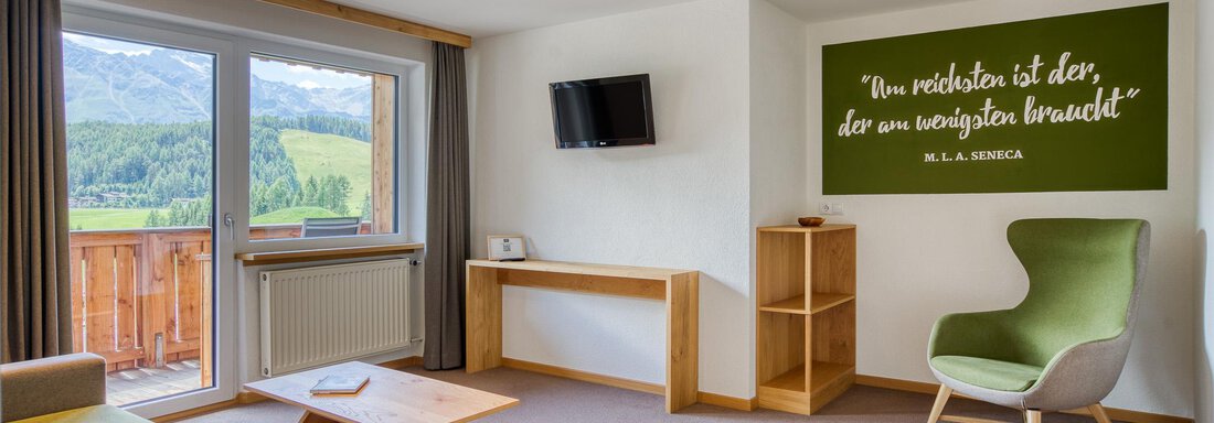 Ein modernes Zimmer mit großen Fenstern und einem Balkon, von dem aus die Landschaft sichtbar ist. In der Ecke steht ein komfortabler Stuhl und an der Wand befindet sich ein inspirierendes Zitat. - Hotel Falknerhof, am Ursprung - Niederthai