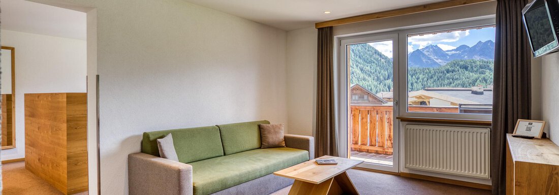 Ein modernes Wohnzimmer mit einem grünen Sofa und einem Holztisch. Große Fenster bieten einen Blick auf die Berge. - Hotel Falknerhof, am Ursprung - Niederthai
