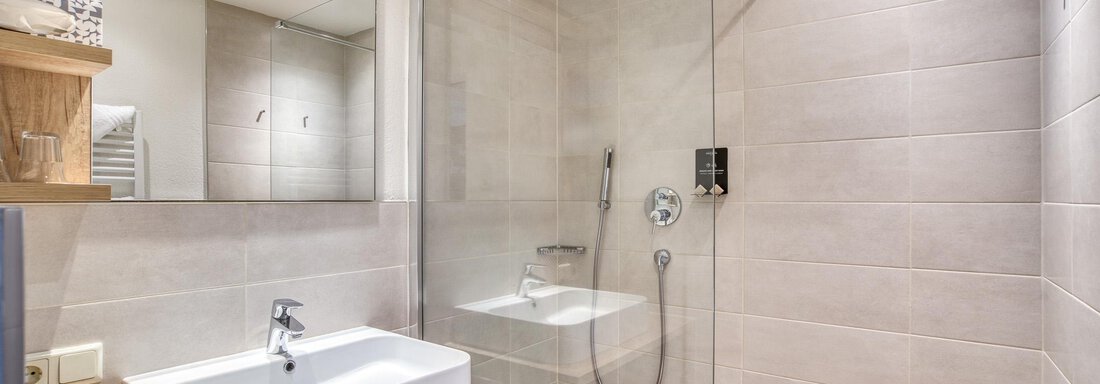 Ein modernes Badezimmer mit einer Dusche und einem eleganten Waschbecken. Die Wände sind hell gefliest und es gibt Holzregale für zusätzliche Ablagefläche. - Hotel Falknerhof, am Ursprung - Niederthai