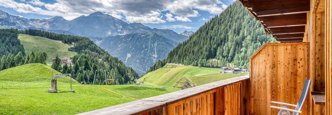 Ein schöner Balkon mit Holzgeländer und einem Stuhl. Im Hintergrund erstrecken sich grüne Hügel und majestätische Berge unter einem blauen Himmel mit Wolken. - Hotel Falknerhof, am Ursprung - Niederthai