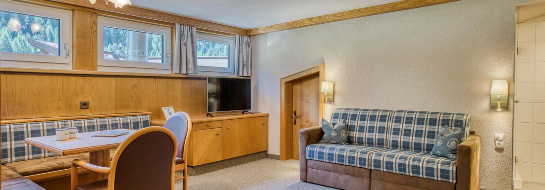 Ein gemütliches Wohnzimmer mit einem Sofa und einem Esstisch. Die Wände sind mit Holz verkleidet und es gibt große Fenster, die viel Licht hereinlassen. - Hotel Falknerhof, am Ursprung - Niederthai