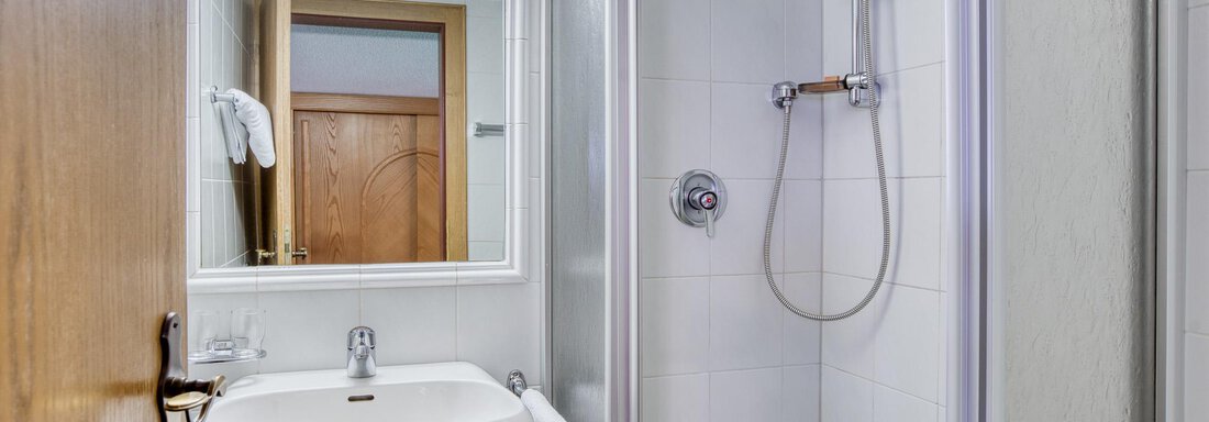 Ein modernes Badezimmer mit einer Dusche, einem Waschbecken und einem Spiegel. Die Wände sind hell gefliest und es gibt einen Holzschrank im Hintergrund. - Hotel Falknerhof, am Ursprung - Niederthai