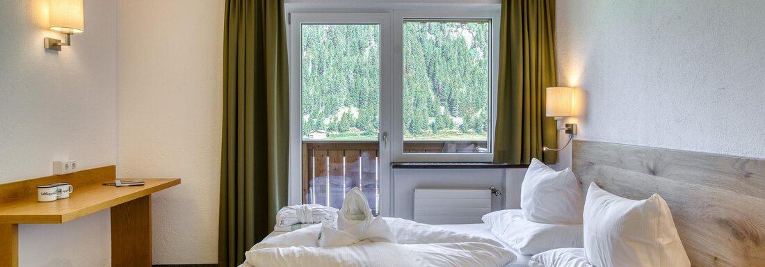 Ein gemütliches Schlafzimmer mit einem großen Bett und zwei Kissen. Durch die Fenster sieht man grüne Bäume und viel Tageslicht. - Hotel Falknerhof, am Ursprung - Niederthai