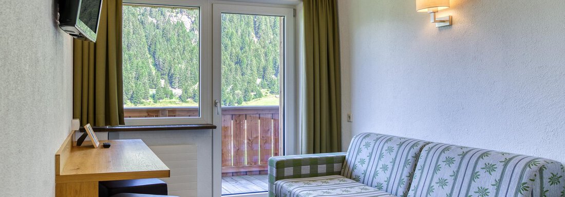 Ein gemütlicher Raum mit einem Sofa und einem Holztisch. Große Fenster bieten einen Blick auf die umgebenden Berge. - Hotel Falknerhof, am Ursprung - Niederthai