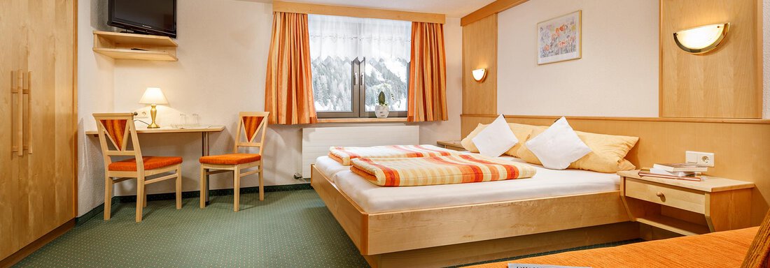 A cozy room with a large bed and orange accents. There is a table, a television, and windows with curtains. - Hotel Tauferberg - Niederthai