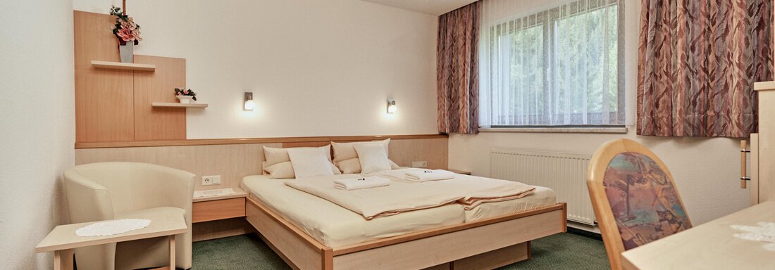 A cozy bedroom with a double bed and green carpet. Next to the bed are a table and a chair as well as a window with curtains. - Hotel Tauferberg - Niederthai