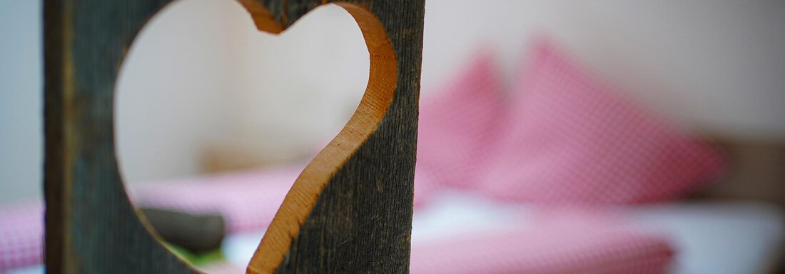 Appartement2_Zimmer0 Ein Holzteil mit einem herzförmigen Ausschnitt. Im Hintergrund sind rote Kissen auf einem Bett zu sehen. - Sonnenhof - Niederthai