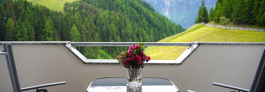 Ein gemütlicher Balkon mit einem Tisch und zwei Stühlen. Im Hintergrund erstrecken sich grüne Hügel und Berge. - Sonnenhof - Niederthai