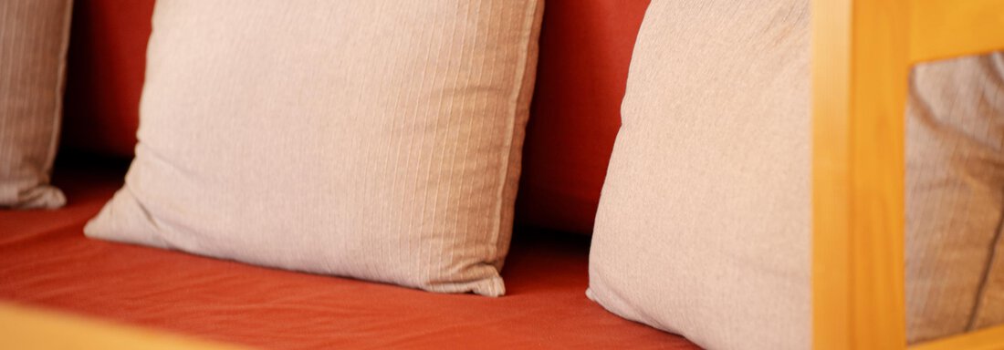 Ein gemütliches Sofa mit zwei hellen Kissen auf rotbraunem Stoff. Die Holzlehne verleiht dem Raum eine warme Atmosphäre. - Sonnenhof - Niederthai