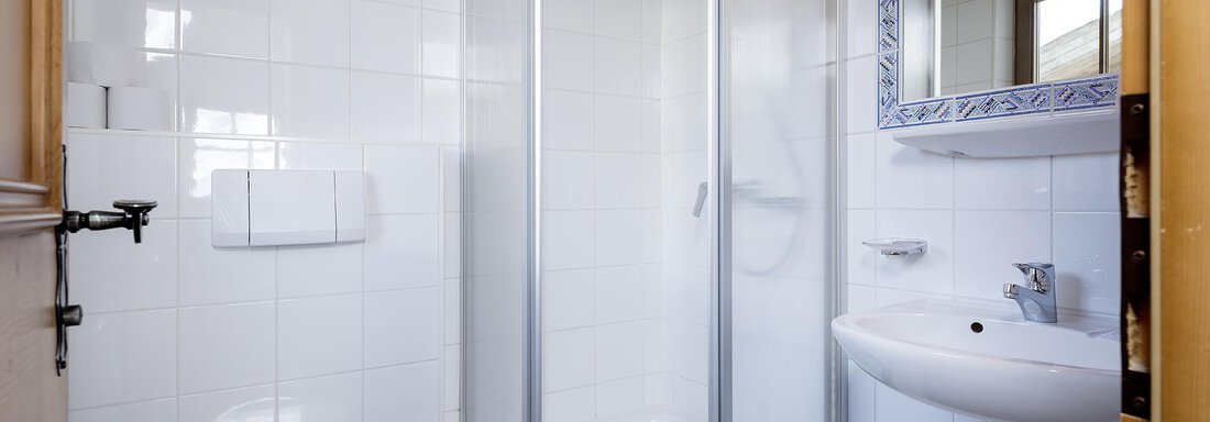 A modern bathroom with a shower and a sink. The walls are tiled in white, and there is a large wall mirror. - Appartementhaus Schalfblick - Obergurgl 