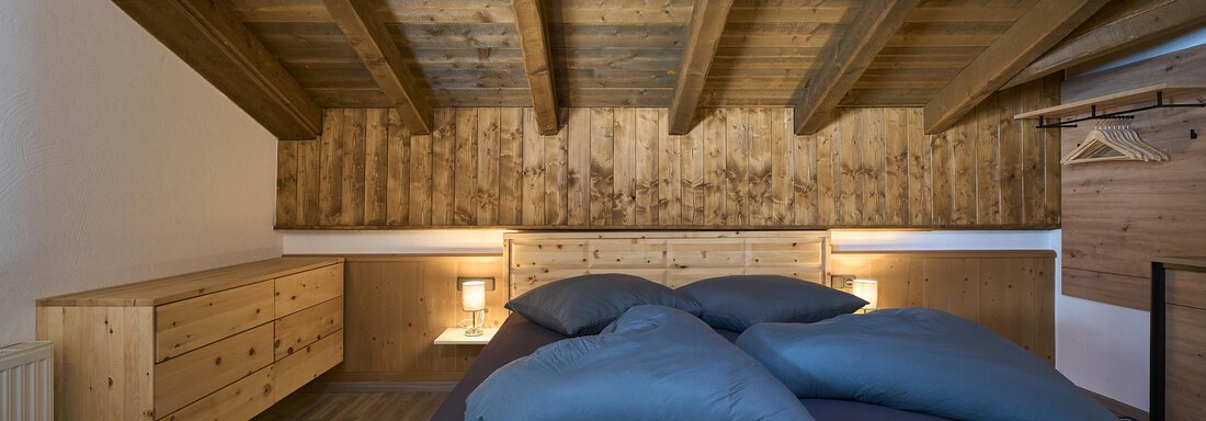 A cozy bedroom with a wooden beam ceiling and modern furniture. The large bed is equipped with gray pillows and a dark bedspread. - Appartementhaus Schalfblick - Obergurgl 