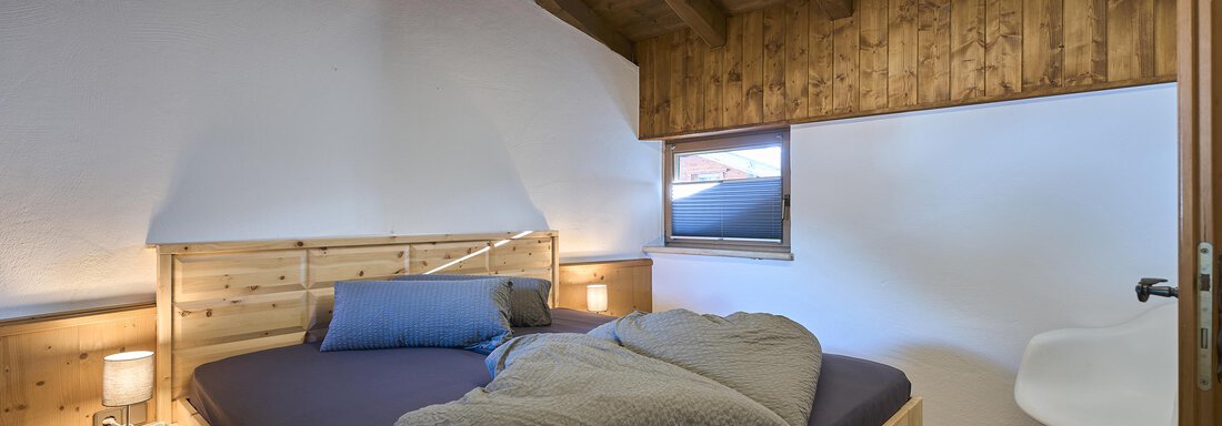 A cozy bedroom with a wooden bed and modern lighting. The walls are paneled with wood, and a window lets in daylight. - Appartementhaus Schalfblick - Obergurgl 