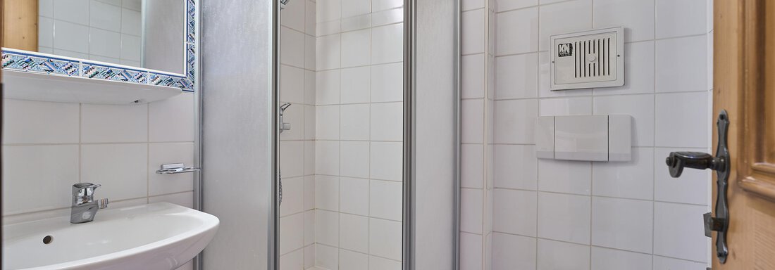A modern bathroom with a shower, sink, and toilet. The walls are tiled white, and the ceiling has wood paneling. - Appartementhaus Schalfblick - Obergurgl 