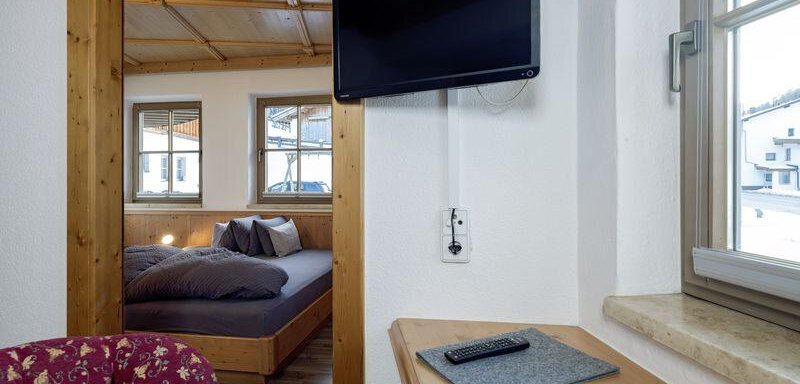 A cozy room with a TV on the wall and a wooden shelf. In the background, there is a double bed and windows that let in natural light. - Appartementhaus Schalfblick - Obergurgl 