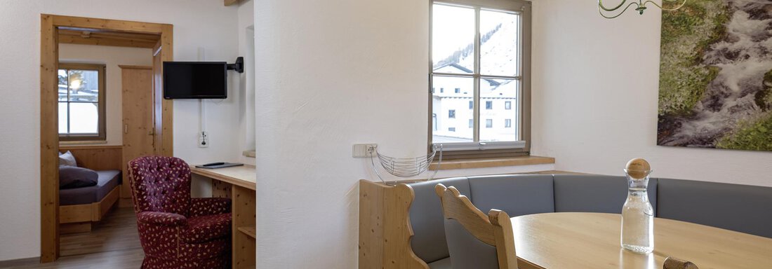 A cozy living space with a dining table, surrounded by a bench and chairs. In the background, a television and another living area are visible. - Appartementhaus Schalfblick - Obergurgl 