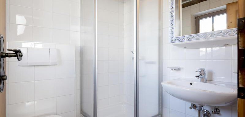 A modern bathroom with a shower, a sink, and a toilet. The walls are light tiled and the light comes in through a window. - Appartementhaus Schalfblick - Obergurgl 