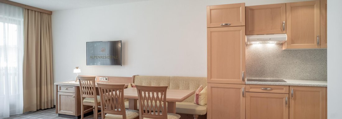 A bright and modern open-plan kitchen with a dining area and a cozy sofa. The furniture is made of light wood and the room offers plenty of light through a large window. - Ferienwohnung de Luxe Alpenresidenz am Mühlbach - Obergurgl 