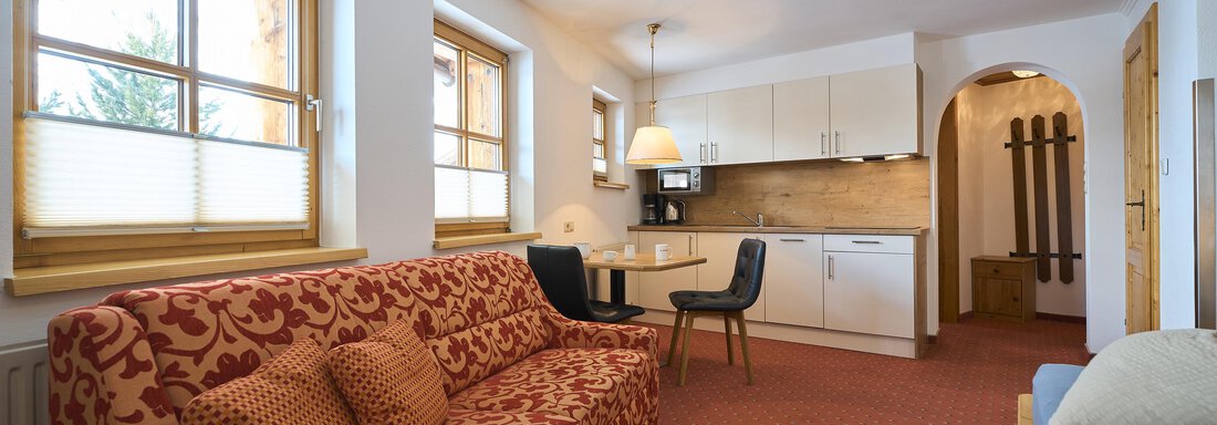 A cozy living room with a red sofa, a table, and chairs. The bright kitchen is visible in the background. - Forest Chalet - Obergurgl 