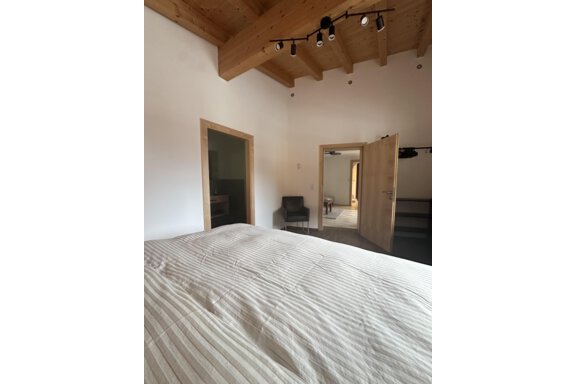 A simple bedroom with a large bed and wooden ceilings. There is a door to another room and a chair in the corner. - Forest Chalet - Obergurgl 