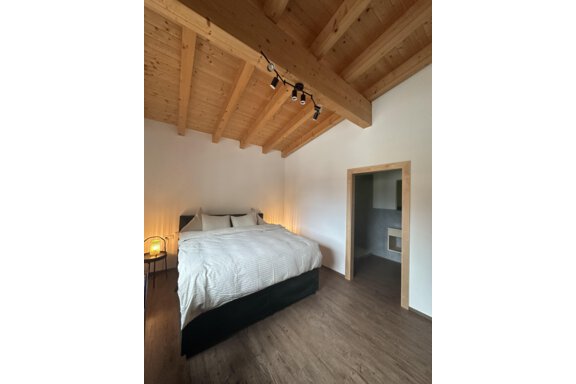 A modern bedroom with a large bed and a wooden ceiling. The walls are painted light, and there is access to another room. - Forest Chalet - Obergurgl 